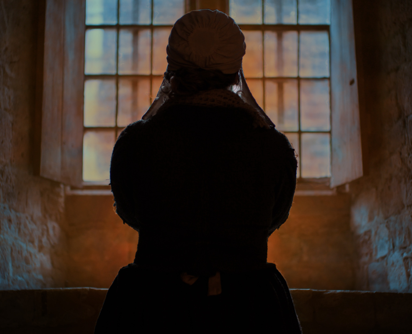 a woman in 17th century dress prays at a window