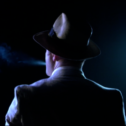 shot from behind, moodily lit and man in a suit and hat exhales smoke